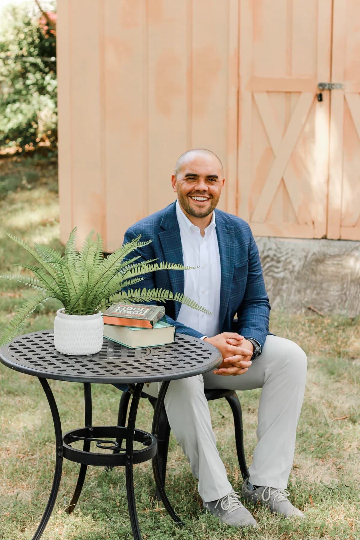Tyson P. Lee, seated outdoors at a small table with books and a fern, smiling.